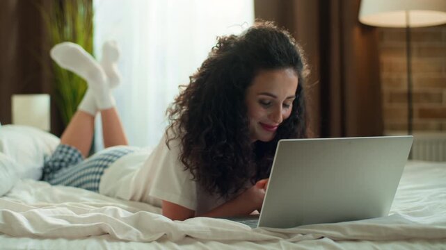 Smiling thinking optimistic Caucasian woman using laptop lying in bed positive gadget addicted female girl with computer e-learning chatting network working scrolling cyberspace at morning in bedroom