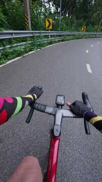First-person point of view of a road cyclist riding through a wet winding mountain road surrounded by lush tropical forest. The footage features roadside guardrails, curve warning signs, and rain-soak