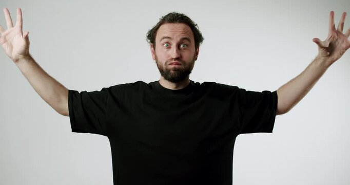 A bearded man in a black t-shirt against a white background expresses surprise and confusion. He throws his hands up in the air, showing his frustration with a situation he cannot understand.