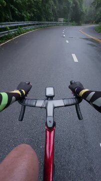 First-person point of view of a road cyclist riding along a wet mountain road after rainfall while passing a runner in a lush tropical forest. The immersive footage highlights endurance sports, outdoo