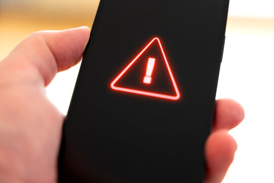 Hand holds a smart phone with a system warning sign on screen, computer crime. Network security, system hacked, firewall, samrtphone virus, data, privacy, safety, danger, mobile phone problems.