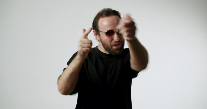 Happy bearded man in sunglasses dances and points at the camera on a white background. He smiles and moves his arms and points his finger while making eye contact. This energetic video conveys joy