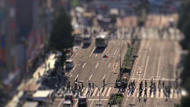 Tilt shift timelapse of vehicles and pedestrians moving through a busy Tokyo city intersection in daylight. Urban traffic flow and crosswalk activity from elevated view.