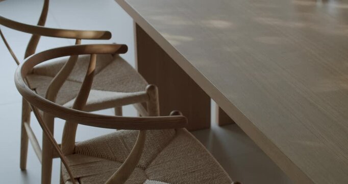 Two empty wooden chairs with woven seats sit beside a long wooden dining room table. Sunlight creates dappled shadows on the table's surface, indicating a bright day. minimalist design atmosphere.