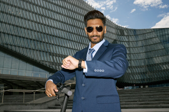 Portrait of young adult Indian businessman checking smartwatch while standing with electric scooter near office building, conveying punctuality and urban mobility. For corporate branding