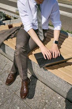 Vertical shot of young adult Caucasian businessman working on laptop while sitting outdoors, managing remote tasks during break. Ideal for hybrid work, mobile productivity marketing