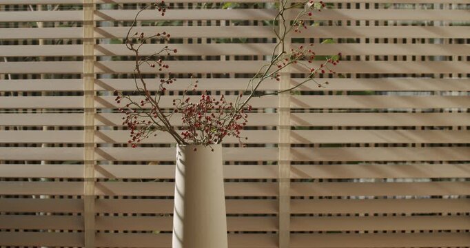 Dried red berries in a white ceramic vase against a minimalist wood slat background. Sunlight casts serene shadows. Perfect for themes of simplicity, nature, and interior design.