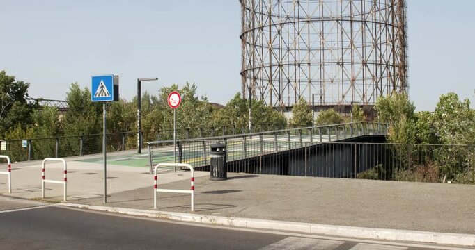 A pedestrian bridge with glass railings leading towards green trees, with road signs, street barriers, and a large metal gasometer structure looming in the background. It's located in Rome, Italy.