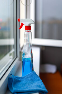 Blue liquid detergent spray bottle and microfiber rag placed on white window sill. Domestic sanitation kit and essential housekeeping tools concept for effective glass surface cleaning.