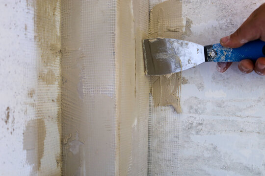 A builder's hand carefully applies plaster mortar with a metal spatula to the inner wall corner protected by a reinforcing mesh. The close-up shot demonstrates the technological process of surface lev