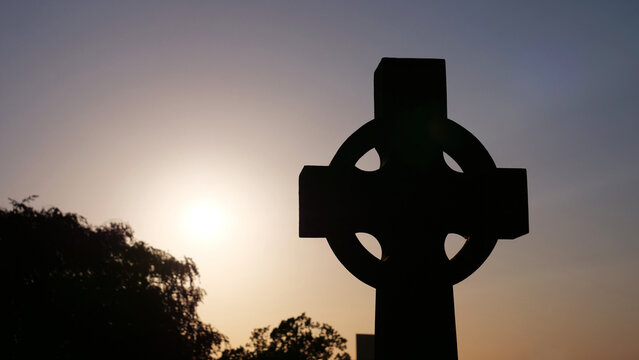 Celtic cross silhouette at sunset, religious stone monument against evening sky, christian spiritual symbol in cemetery graveyard at dawn