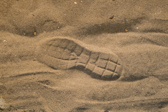 Close-up of a boot footprint impressed in textured brown beach sand. Natural surface detail. Human presence, travel, coastal environment, and outdoor exploration concept