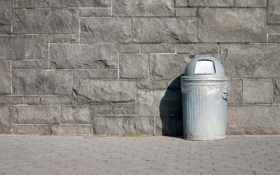 Metal trash bin on NYC sidewalk. Outdoor public waste receptacle sitting beside stone wall in sunlight.