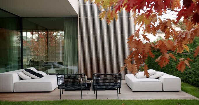 Modern house with garden furniture on wooden deck in backyard. Cozy white sofas and black wicker chairs. Stylish exterior design of contemporary home in autumn. Red oak leaves on branches.