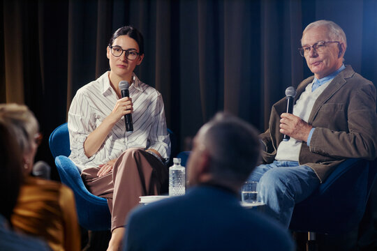 Caucasian young woman and senior man speaking into microphones during panel discussion on stage, sharing expertise and leadership. Ideal for business events, conferences, PR