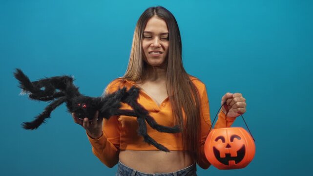 Woman holding orange pumpkin bucket and large fuzzy spider with outstretched hands against studio backdrop; festive fun.
