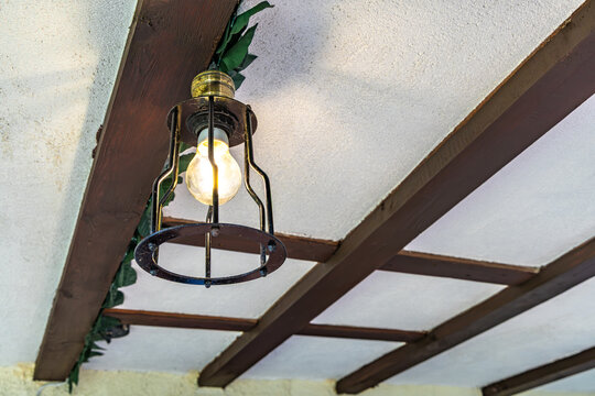 A man replaces a burned-out light bulb in a fixture on a porch in front of a house in the garden, unscrewing and screwing in a burned-out LED bulb in a lamp hanging from the ceiling 