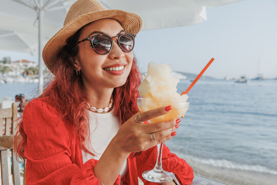 Happy woman wearing a hat and sunglasses, sitting at a beach bar enjoying a refreshing frozen drink during summer vacation