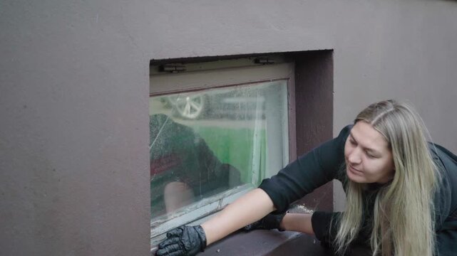 Woman in work gloves inspects a dirty, damaged basement window frame, considering home repair or replacement.
