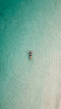 Overhead view of woman floating in shallow turquoise water with concentric ripples