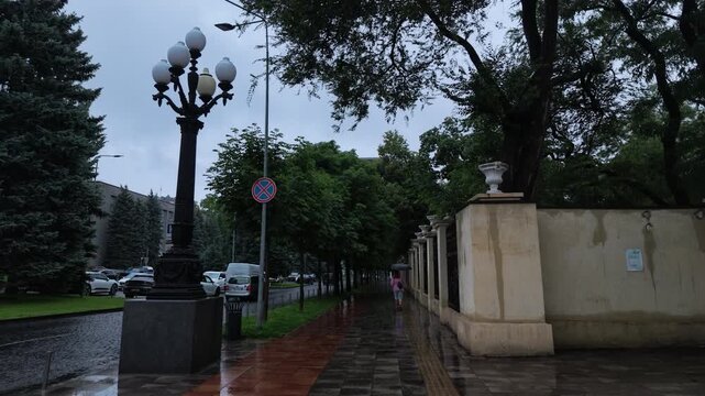 A lonely pedestrian with a pink umbrella walks on a wet sidewalk along a vintage street lamp and historical wall during rain. Overcast day, moody urban atmosphere, water reflections.