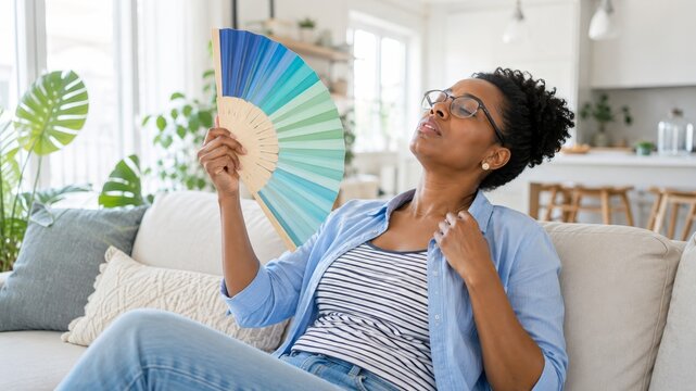 Woman experiencing menopause hot flash while cooling with hand fan at home, hormonal change, midlife health, sudden heat, sweating discomfort, indoor warmth, self care during climacteric