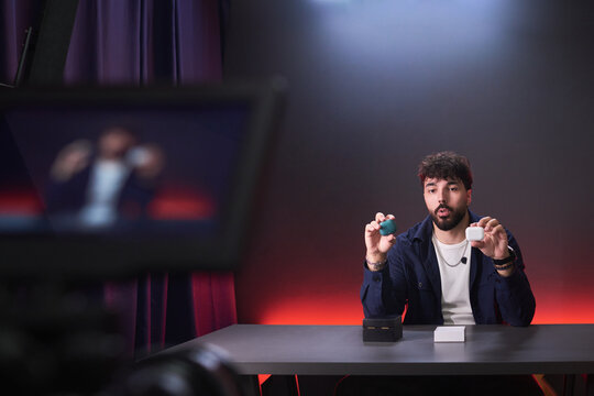 Young man presenting wireless earbuds cases on camera while recording product review at desk, comparing features for tech content. Ideal for influencer marketing, ecommerce promotion