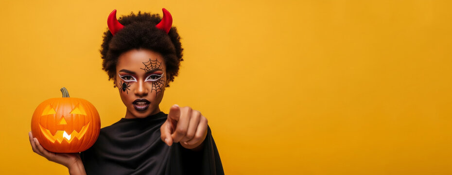 African American woman dressed as a devil holding a glowing pumpkin lantern and pointing at camera. Halloween celebration character in creative makeup. Holiday costume party concept.
