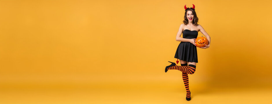 Woman dressed in devil costume holding halloween pumpkin. Girl in black party outfit with horns and striped stocking posing on orange background. Festive celebration concept, copy space.