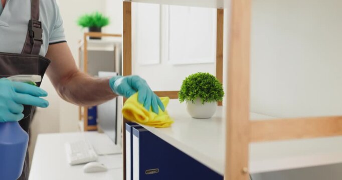 Cleaner team disinfecting office workplace wiping surfaces. Gloved janitor uses spray and cloth to sanitize a shelf and tidy desks in a modern corporate office. Clear workplace hygiene concept.