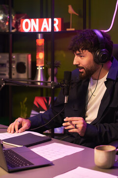 Vertical shot of young Caucasian man hosting podcast, speaking into microphone with headphones at desk. Suitable for audio media, streaming, content marketing campaigns