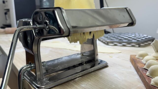 Stainless steel pasta machine on a floured wooden counter with fresh dough sheets and cut pieces nearby, cozy home kitchen scene, conveys handmade cooking and preparation