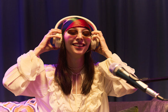 Young woman adjusting headphones while recording audio near microphone, smiling during podcast or music session. Useful for creator economy, streaming, entertainment marketing