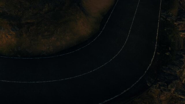 A winding road can be seen cutting through rocky terrain during dusk. The dark sky adds a dramatic backdrop to the scene while shadows play on the ground.