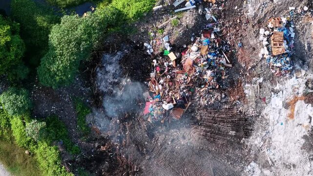 Smoke rises from burning trash piles. Green trees border the smoky waste