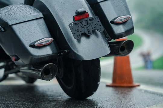 Close up of motorcycle rear end in rain, showcasing wet surfaces and traffic cone on wet road