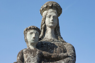 Obraz premium Weathered stone statue of the crowned Virgin Mary holding the Christ Child beneath a clear blue sky.