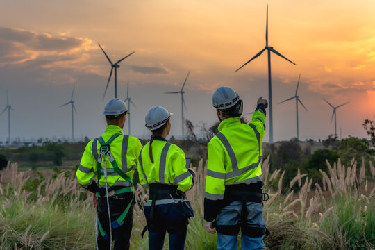 Rear view team of technicians working until dusk, conducting maintenance checks on a wind turbine generator surrounded by farmlands at twilight