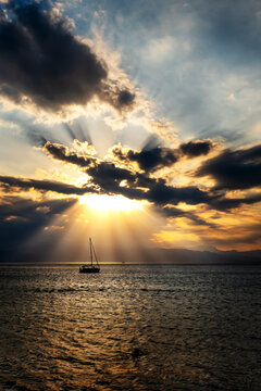 Silhouetted Sailboat at Golden Sunset Over Ionian Sea with dramatic piercing Sunrays. Breathtaking view of a vivid golden-orange sunset over the calm Ionian Sea, featuring a silhouette of a sailboat g