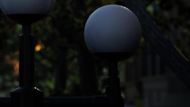 Light fixtures are positioned in a park as evening approaches. The setting sun casts shadows and highlights the round shapes of the lights. Trees fill the background.