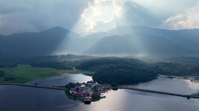 Morning sun rays break through clouds illuminating a tranquil lake surrounded by forest and distant mountains creating a serene at Tasik Raban