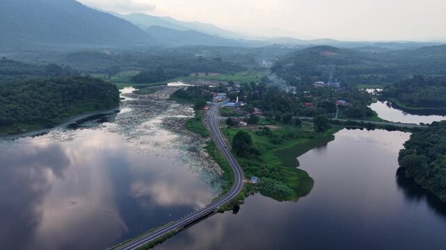 Aerial view captures a winding road beside a calm lake. Mountains reflect in the water's surface at Tasik Raban