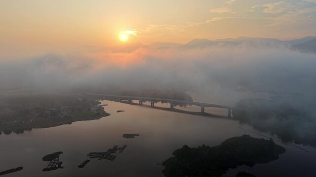 A misty sunrise casts soft golden light over a calm river with a bridge spanning across foggy cloud at Tasik Raban