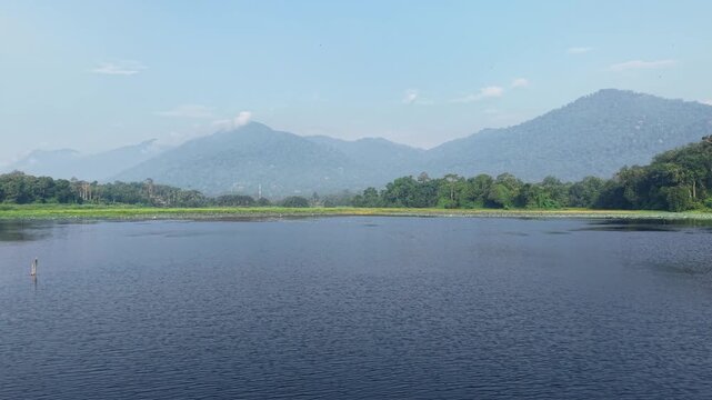 A tranquil lake with still water reflecting dense green forest and a distant misty mountain under a clear blue sky on a peaceful daytime at Tasik Raban