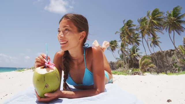 Beach bikini woman relaxing drinking fresh coconut water lying down sunbathing on fun Caribbean vacation tanning. Asian model enjoying suntan. Real unretouched woman model without any makeup