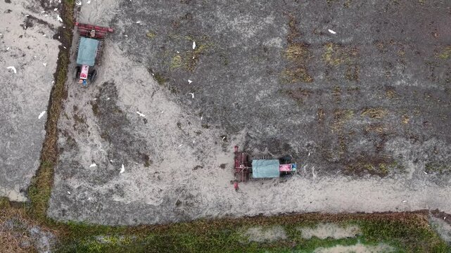 Aerial view shows two tractors working in a muddy field. White egret birds fly above the machinery