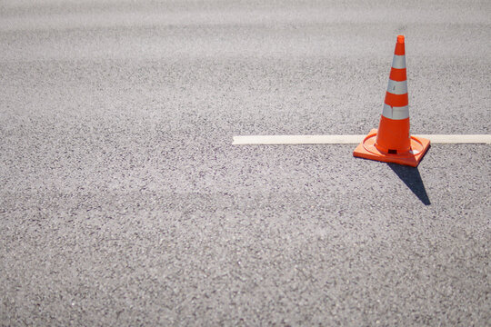 Orange traffic cone with white reflective stripes on a textured gray asphalt road. Minimalist composition with selective focus and distinct shadows on the pavement.