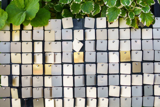 Square metal sequins on a matrix panel combined with green plants at the top. Shimmering silver and gold metallic texture with natural variegated leaves.