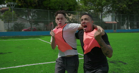 Fototapeta premium Latin and White teammates celebrate goal on urban soccer field, running with arms in air, pumping fists and shouting, embracing and smiling, showing unity and triumph