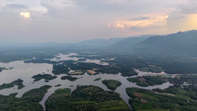 Aerial panorama showing a river winding through green islands and forested terrain with mountain range in the background under a cloudy sunset sky at Tasik Raban